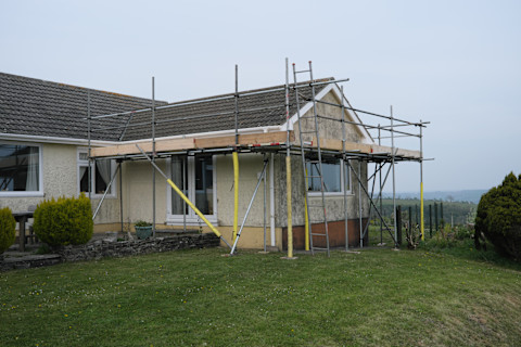Scaffolding erected around the extension on a bungalow, ready to commence work on a roof repair. The house is set in a grassy garden with bushes. There is an overcast sky behind it, and a foggy view of rolling hills.