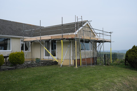 Scaffolding erected around the extension on a bungalow, ready to commence work on a roof repair. The house is set in a grassy garden with bushes. There is an overcast sky behind it, and a foggy view of rolling hills.